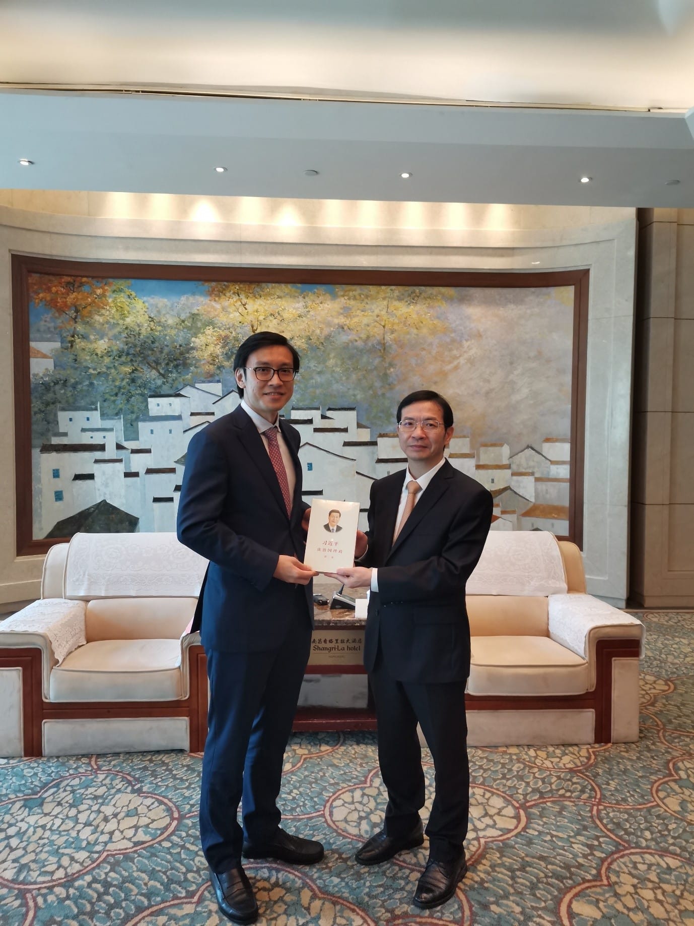 Two men in suits exchange a book with a portrait of Xi Jinping, in Shangri-La Hotel.